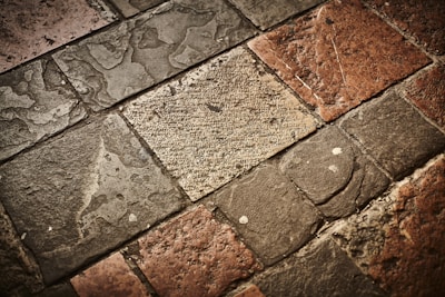 A textured surface composed of irregularly shaped stone tiles in various shades of brown, grey, and beige. The stones have a worn appearance with visible imperfections and some patina.