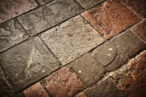 A textured surface composed of irregularly shaped stone tiles in various shades of brown, grey, and beige. The stones have a worn appearance with visible imperfections and some patina.