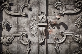 Intricate wrought ironwork with ornate floral and scroll patterns in a symmetrical design on an aged, textured metallic surface.