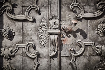 Intricate wrought ironwork with ornate floral and scroll patterns in a symmetrical design on an aged, textured metallic surface.