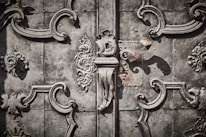 Intricate wrought ironwork with ornate floral and scroll patterns in a symmetrical design on an aged, textured metallic surface.