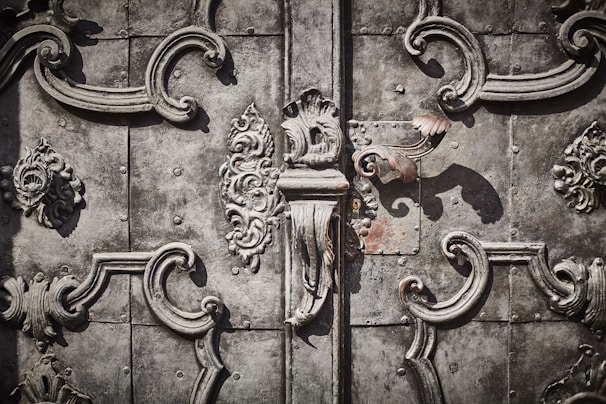 Intricate wrought ironwork with ornate floral and scroll patterns in a symmetrical design on an aged, textured metallic surface.