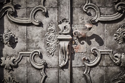 Intricate wrought ironwork with ornate floral and scroll patterns in a symmetrical design on an aged, textured metallic surface.