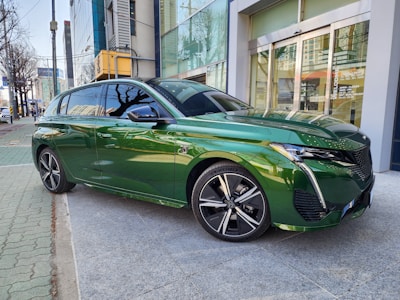 A sleek, green car is parked on a sidewalk in an urban area. The modern design of the car includes bold lines and a shiny exterior. Surrounding buildings and trees are reflected on the car's surface, enhancing its glossy finish. Background architecture suggests a mix of commercial and residential buildings, with a clear sky above.