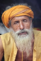 An elderly man with a long white beard and mustache wears a bright orange turban and yellow shirt, displaying a serene and contemplative expression. The background is dark, highlighting his facial features and attire.