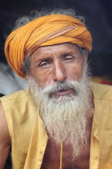 An elderly man with a long white beard and mustache wears a bright orange turban and yellow shirt, displaying a serene and contemplative expression. The background is dark, highlighting his facial features and attire.