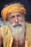 An elderly man with a long white beard and mustache wears a bright orange turban and yellow shirt, displaying a serene and contemplative expression. The background is dark, highlighting his facial features and attire.