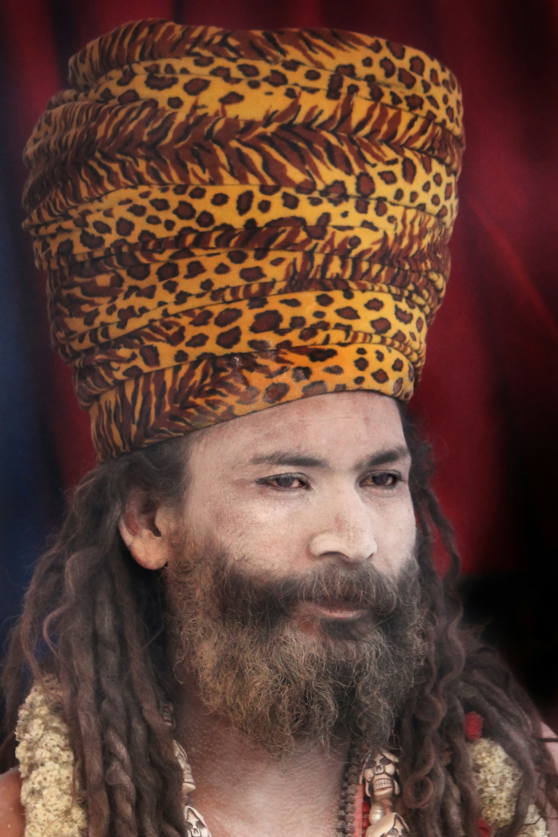A person with long dreadlocks and a bushy beard wears a tall, patterned headdress resembling leopard print. The expression appears contemplative, and the background is a deep red with some indistinct shadows.