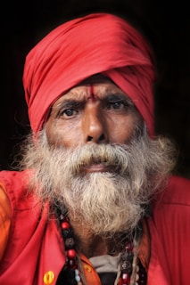 An elderly man with a long white beard is wearing a red turban and robe. Beads hang from his neck, and his expression is serious and contemplative. Natural light highlights the textures of his skin and fabric.