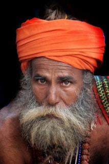 An elderly man with a long, full beard wears an orange turban and a colorful cloth draped over his shoulder. His expression is contemplative and serene, framed by his deeply set eyes. The vibrant colors of his clothing contrast with the dark background, highlighting his facial features and garments.