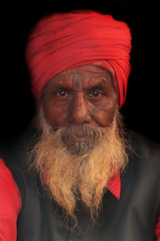 An elderly man with a long white beard, wearing a vibrant red turban and matching red sleeves. His expression is serious and contemplative, set against a dark background that emphasizes his features.