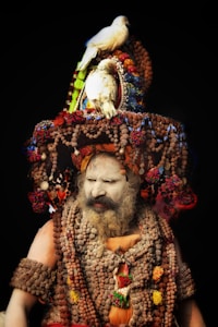 A person is wearing elaborate traditional attire adorned with numerous wooden beads and colorful decorations. The face appears to be painted white, with a thick beard visible. There are two white birds perched on top of the ornate headdress, which is covered with intricate beadwork and patterns.