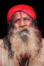A man with a long, thick white beard and a mustache is wearing a bright red turban and has a red mark on his forehead. He is dressed in a beige garment against a dark background.