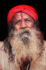 A man with a long, thick white beard and a mustache is wearing a bright red turban and has a red mark on his forehead. He is dressed in a beige garment against a dark background.
