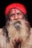 A man with a long, thick white beard and a mustache is wearing a bright red turban and has a red mark on his forehead. He is dressed in a beige garment against a dark background.