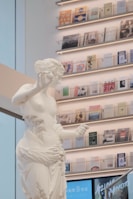 A white marble statue depicting a classical figure is positioned in front of a wall with neatly arranged shelves filled with various books and magazines. The shelving features soft lighting that enhances the overall modern and artistic atmosphere.