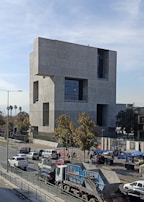 A large, modern concrete building with a geometric design dominates the scene. It is surrounded by a busy urban environment with several vehicles, including cars and a dump truck, moving along the street. Trees with leaves add some greenery to the area, while people can be seen walking near the building.