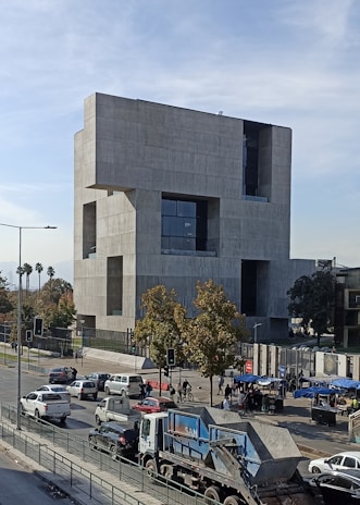 A large, modern concrete building with a geometric design dominates the scene. It is surrounded by a busy urban environment with several vehicles, including cars and a dump truck, moving along the street. Trees with leaves add some greenery to the area, while people can be seen walking near the building.
