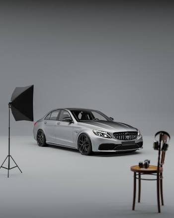 A sleek silver car is prominently positioned in a studio setting, featuring a softbox lighting setup on a stand to the left. In the foreground sits a simple wooden chair with a camera and a pair of headphones resting on top.