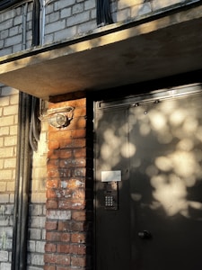 A dimly lit exterior of a brick building features a door with a keypad entry system. Shadows of leaves create patterns on the surface of the door, and various pipes run along the brick wall.