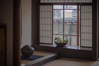 A serene interior of a traditional Japanese room with a shoji screen window casting soft light. A small basket with green plants is placed on the windowsill. A ceramic pot rests on a low table covered with a blue cloth.