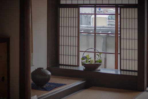 A serene interior of a traditional Japanese room with a shoji screen window casting soft light. A small basket with green plants is placed on the windowsill. A ceramic pot rests on a low table covered with a blue cloth.
