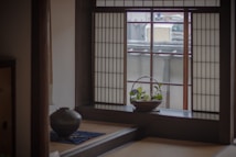 A serene interior of a traditional Japanese room with a shoji screen window casting soft light. A small basket with green plants is placed on the windowsill. A ceramic pot rests on a low table covered with a blue cloth.