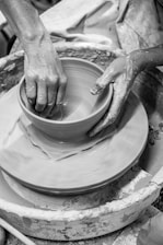 Hands shape a clay pot on a rotating pottery wheel, with wet clay and splatters visible around. The focus is on the tactile interaction with the clay, highlighting the craft and artistry involved.