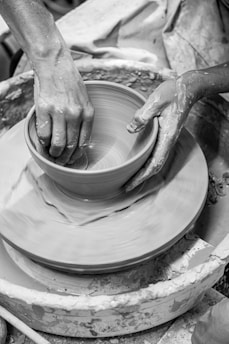 Hands shape a clay pot on a rotating pottery wheel, with wet clay and splatters visible around. The focus is on the tactile interaction with the clay, highlighting the craft and artistry involved.