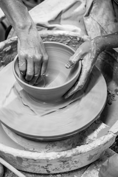 Hands shape a clay pot on a rotating pottery wheel, with wet clay and splatters visible around. The focus is on the tactile interaction with the clay, highlighting the craft and artistry involved.