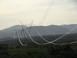 A series of electrical power lines stretch across a rural landscape with hills in the background. The power lines are supported by tall metal towers that are evenly spaced across the green, forested terrain. The sky is overcast, casting a muted light over the scene.