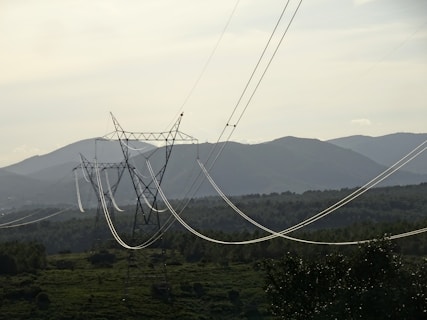 A series of electrical power lines stretch across a rural landscape with hills in the background. The power lines are supported by tall metal towers that are evenly spaced across the green, forested terrain. The sky is overcast, casting a muted light over the scene.