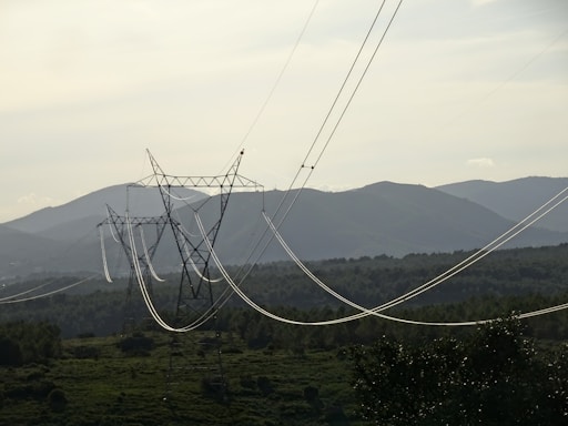 A series of electrical power lines stretch across a rural landscape with hills in the background. The power lines are supported by tall metal towers that are evenly spaced across the green, forested terrain. The sky is overcast, casting a muted light over the scene.