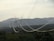 A series of electrical power lines stretch across a rural landscape with hills in the background. The power lines are supported by tall metal towers that are evenly spaced across the green, forested terrain. The sky is overcast, casting a muted light over the scene.