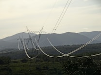A series of electrical power lines stretch across a rural landscape with hills in the background. The power lines are supported by tall metal towers that are evenly spaced across the green, forested terrain. The sky is overcast, casting a muted light over the scene.