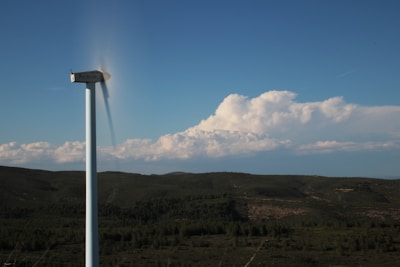 A single wind turbine stands tall in a vast green landscape with rolling hills. Its blades are spinning rapidly, causing a motion blur effect. In the background, there is a clear blue sky with a large formation of white fluffy clouds along the horizon.