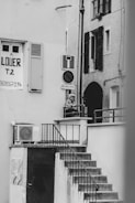 A black and white photograph captures an outdoor staircase leading up to a small landing area, where a person is leaning on the railing. A 'for rent' sign is visible on one of the building walls, along with various signs such as a no entry sign. The architecture features shuttered windows and an exterior air conditioning unit.