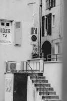 A black and white photograph captures an outdoor staircase leading up to a small landing area, where a person is leaning on the railing. A 'for rent' sign is visible on one of the building walls, along with various signs such as a no entry sign. The architecture features shuttered windows and an exterior air conditioning unit.