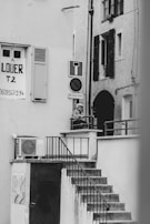 A black and white photograph captures an outdoor staircase leading up to a small landing area, where a person is leaning on the railing. A 'for rent' sign is visible on one of the building walls, along with various signs such as a no entry sign. The architecture features shuttered windows and an exterior air conditioning unit.