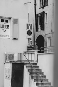 A black and white photograph captures an outdoor staircase leading up to a small landing area, where a person is leaning on the railing. A 'for rent' sign is visible on one of the building walls, along with various signs such as a no entry sign. The architecture features shuttered windows and an exterior air conditioning unit.