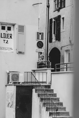 A black and white photograph captures an outdoor staircase leading up to a small landing area, where a person is leaning on the railing. A 'for rent' sign is visible on one of the building walls, along with various signs such as a no entry sign. The architecture features shuttered windows and an exterior air conditioning unit.