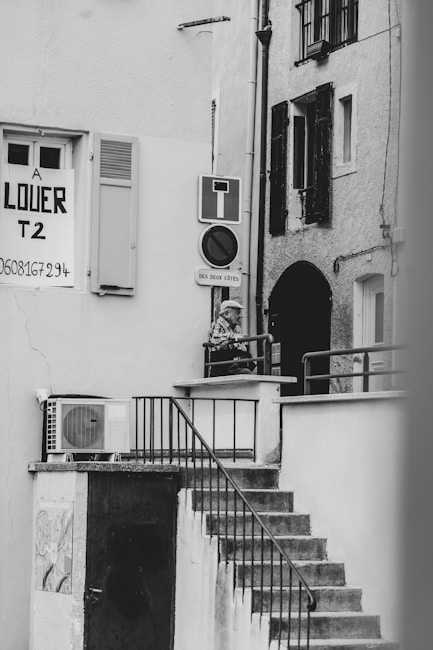 A black and white photograph captures an outdoor staircase leading up to a small landing area, where a person is leaning on the railing. A 'for rent' sign is visible on one of the building walls, along with various signs such as a no entry sign. The architecture features shuttered windows and an exterior air conditioning unit.