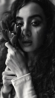 A woman with curly hair is holding a bouquet of flowers close to her face. Her expression is contemplative and a single tear is visible on her cheek. The image is in black and white, emphasizing shadows and light.