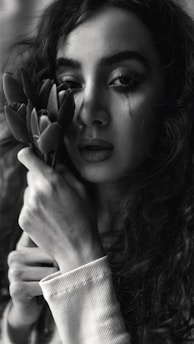 A woman with curly hair is holding a bouquet of flowers close to her face. Her expression is contemplative and a single tear is visible on her cheek. The image is in black and white, emphasizing shadows and light.