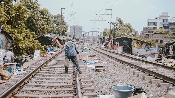 A person wearing a hooded jacket is walking along railway tracks in an urban area. Simple shelters made of tarps and other materials are set up on both sides of the tracks. There are various items and clothing spread on the ground, possibly for sale. Leafy trees and some multi-story buildings are visible in the background, while overhead wires run above the tracks.
