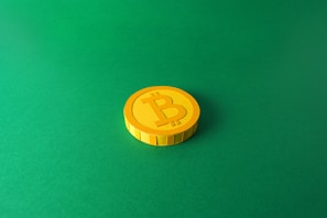 A single stylized yellow coin with a Bitcoin symbol is placed on a solid green background. The coin is centered towards the right with distinct shadows giving it a three-dimensional appearance.