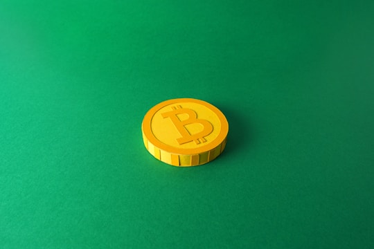 A single stylized yellow coin with a Bitcoin symbol is placed on a solid green background. The coin is centered towards the right with distinct shadows giving it a three-dimensional appearance.