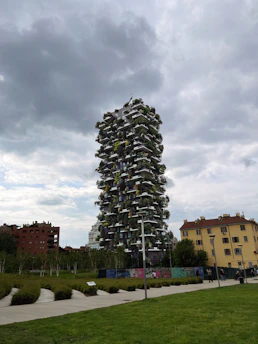 A modern skyscraper covered with vegetation stands amidst a surrounding urban environment. The sky is overcast, creating a contrast with the vivid greenery on the building. Nearby, various structures and trees complement the innovative design.
