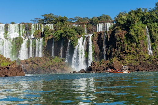 A stunning view of a large waterfall cascading over a series of rocky cliffs surrounded by lush green vegetation. Below, a boat filled with people approaches the base of the falls, creating a sense of adventure.