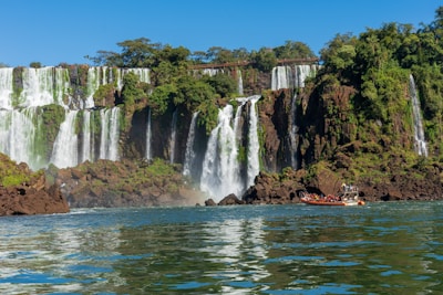 A stunning view of a large waterfall cascading over a series of rocky cliffs surrounded by lush green vegetation. Below, a boat filled with people approaches the base of the falls, creating a sense of adventure.